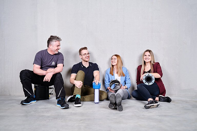 Mitarbeiter sitzen lachend auf dem Boden. Jeder hält ein kleines Produkt von Hennlich in der Hand